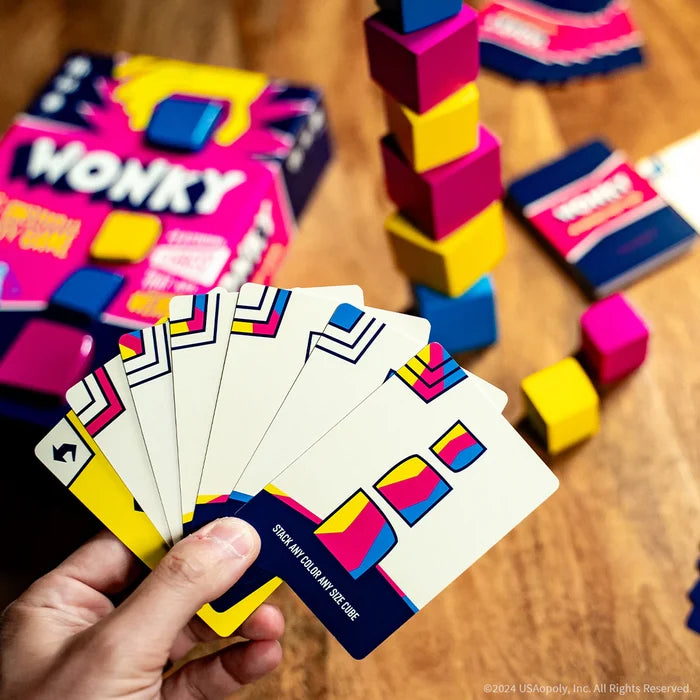 Hand holding colorful game cards in front of a stack of pink, yellow, and blue irregular blocks and a bright game box.