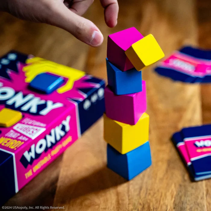 Hand balancing a yellow cube on a tower of blue and pink blocks beside a colorful game box and cards.