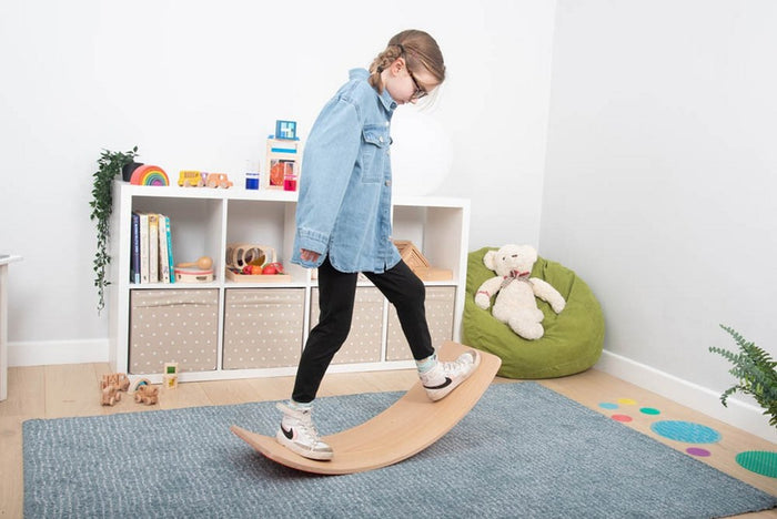 Child balancing on a curved, light wood balance board on a blue rug in a bright playroom with toys.