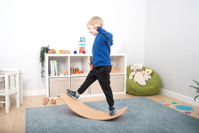Child balancing on a curved natural wood rocker board in a bright playroom with toys and a blue rug.