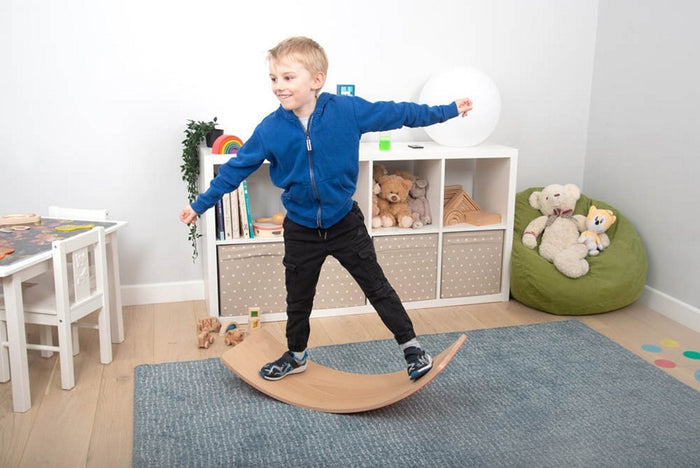 Child balancing with arms outstretched on a curved, natural wood board over a blue rug.