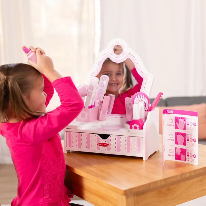 Young girl playing with a pink and white wooden toy vanity with mirror, drawer, and hair and nail accessories.