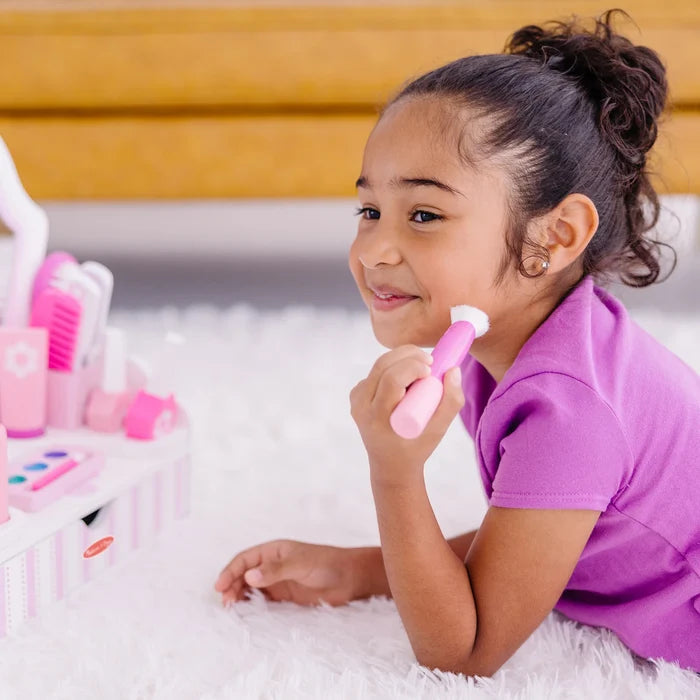 Young girl using a pink toy makeup brush from a striped wooden vanity set while lying on a white rug.