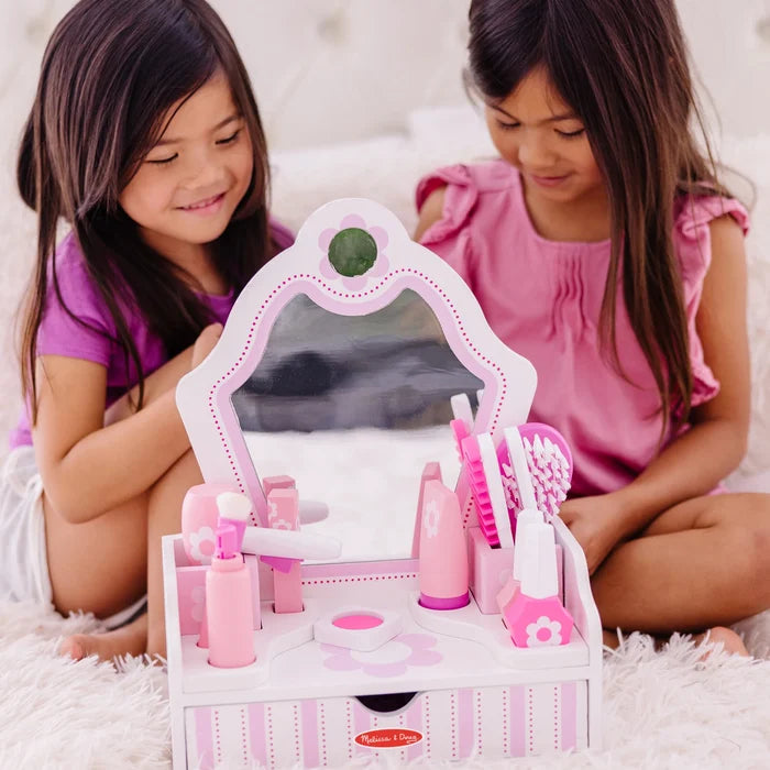 Two girls play with a pink and white wooden toy vanity featuring a mirror, floral accents, and beauty accessories.