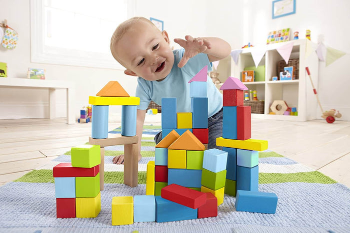 Toddler reaching for a tall tower of multicolor wooden blocks in shapes like cubes and triangles on a striped rug.