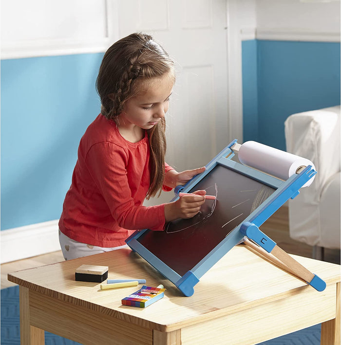 Child using a blue wooden tabletop easel with a chalkboard, paper roll, and chalk set on a light wood table.