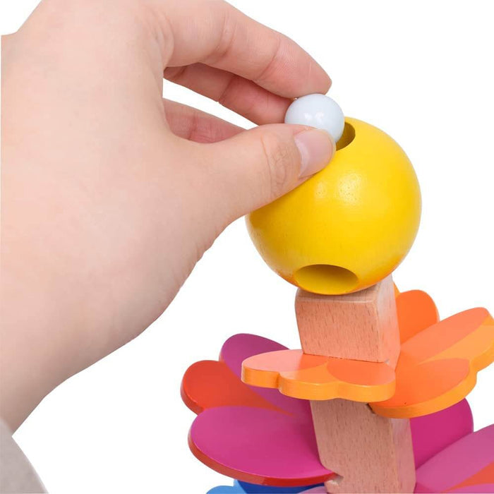 A hand placing a white marble into a yellow sphere atop a colorful wooden toy with flower-shaped levels.