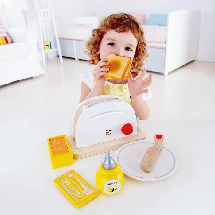 Child playing with wooden white toaster, toy toast, plate, wooden knife, and pretend honey and butter.