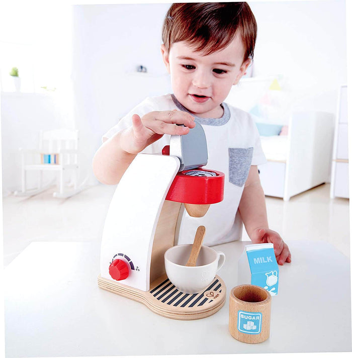 Child playing with a white and wooden toy coffee maker with a red dial, cup, blue milk carton, and wooden sugar jar.