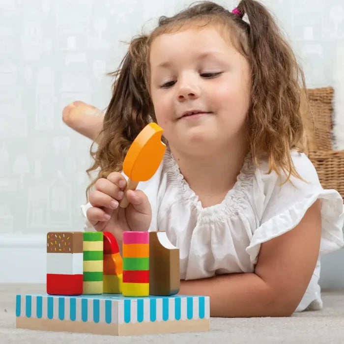 Young girl holding an orange wooden popsicle toy with colorful, striped ice lollies on a blue-striped stand.