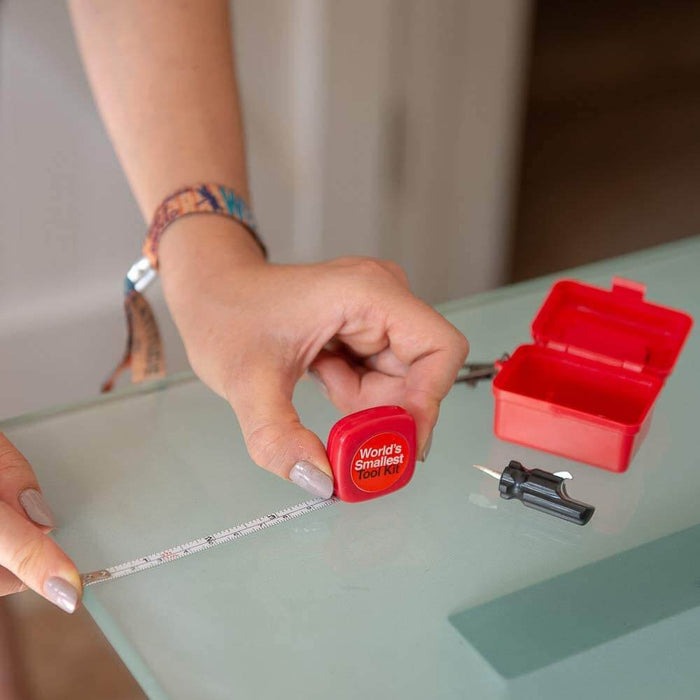 Fingers holding a miniature red tape measure next to an open red toolbox and a small black screwdriver.