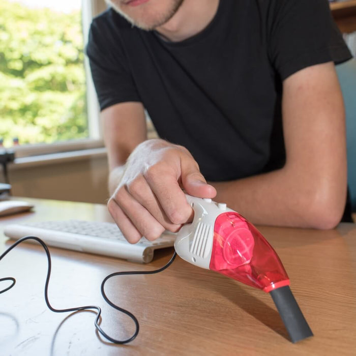 Hand holding a miniature red and white handheld vacuum with a black nozzle and cord over a wooden desk.