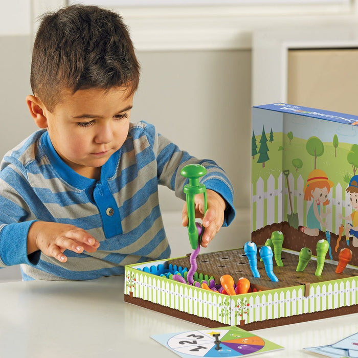 Young boy using green tongs to pull colorful worms from a garden game board box with a spinner nearby.