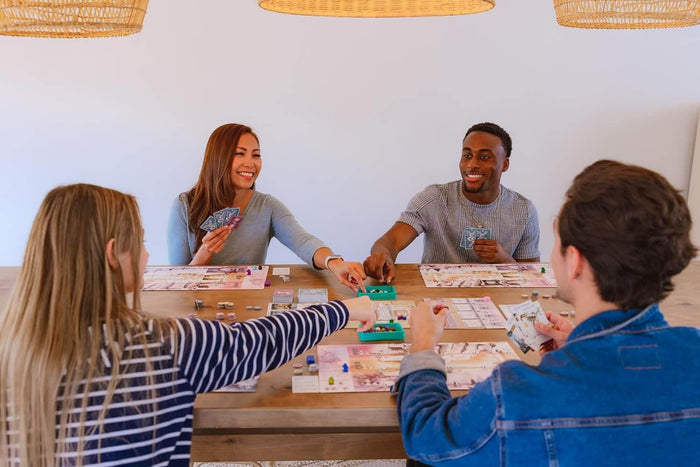 Four people play Wyrmspan board game with illustrated player mats, colorful cards, and tokens on a wooden table.