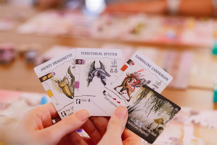 Hand holding three cards featuring colorful dragon illustrations and game icons from the Wyrmspan board game.