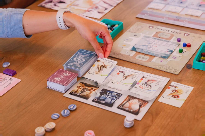 Hand placing a dragon card onto a game board with colorful wooden tokens, egg-shaped pieces, and illustrated cards.