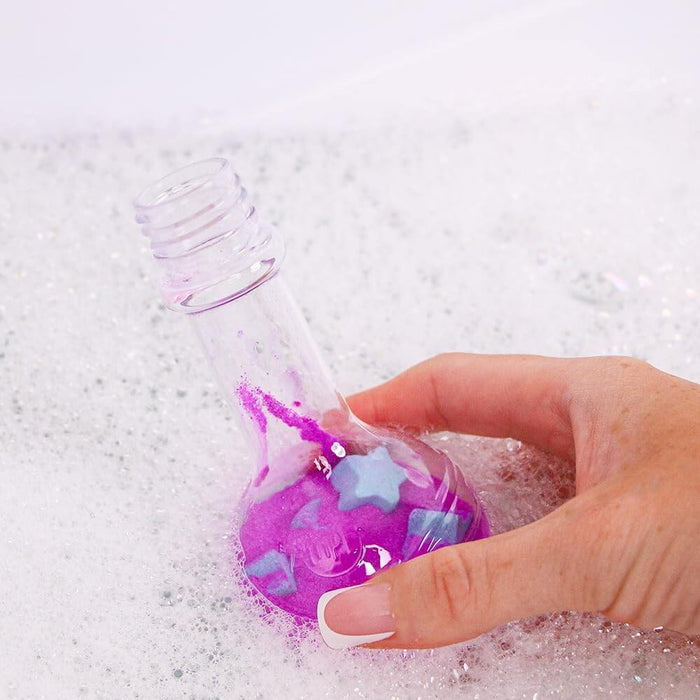 Hand holding a clear flask with purple fizzing powder and blue stars over a bubbly white bath.