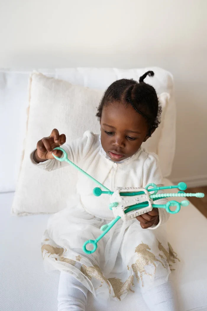 Toddler pulls a textured mint green silicone string on a white sensory activity toy.