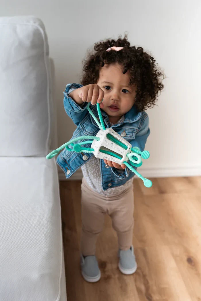 Toddler holds a white speckled activity toy with several flexible, textured teal pull strings and rounded ends.