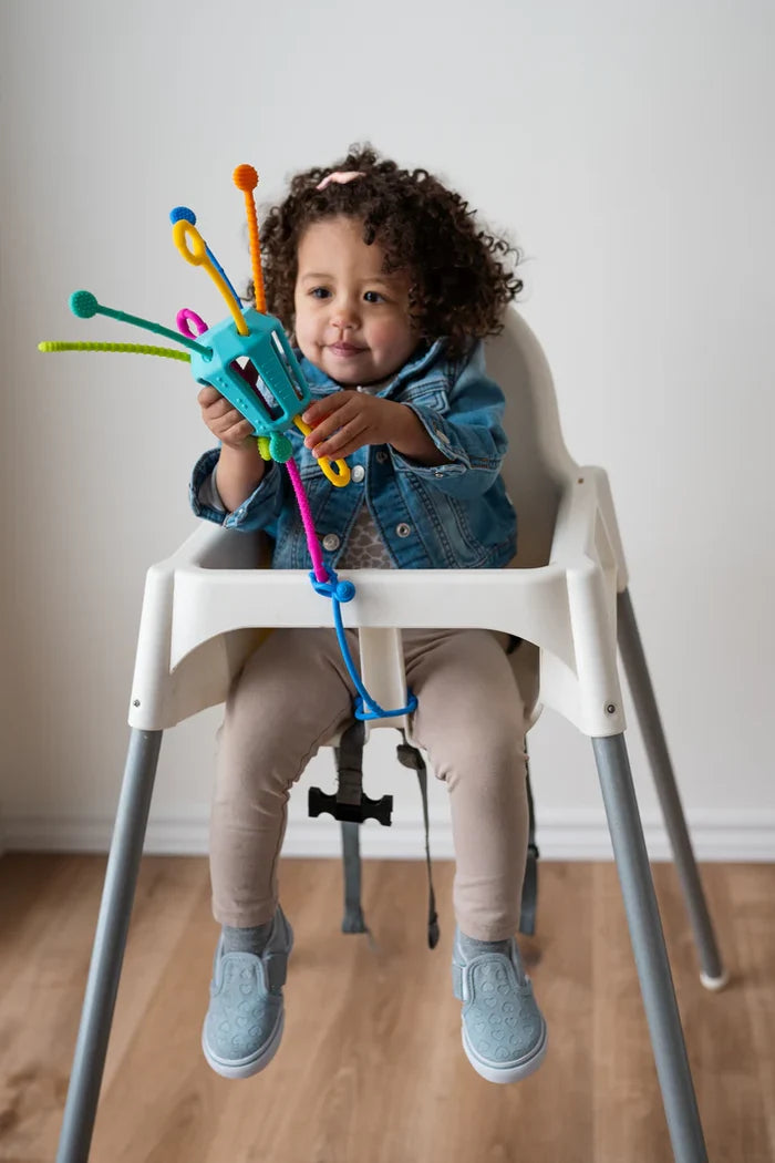 Toddler in a high chair holding a teal silicone sensory toy with colorful, textured pull strings.