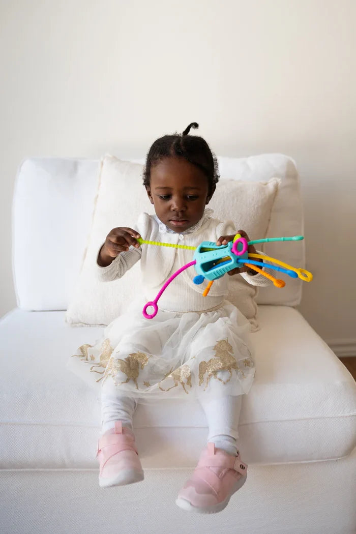 Toddler on a white chair playing with a blue silicone toy featuring vibrant, textured cords in multiple colors.